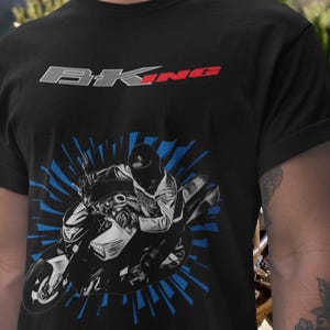 May include: Black t-shirt featuring a motorcycle graphic with a rider. The design includes the text "BKNG" in gray and red. A blue burst pattern surrounds the motorcycle illustration. The shirt is made of a dark fabric.