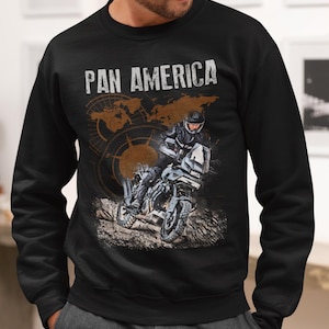 Könnte beinhalten: Schwarzes Sweatshirt mit dem weißen Schriftzug "PAN AMERICA" über einer Grafik eines Motorrads und Fahrers. Das Design beinhaltet eine Karte und einen Kompass in Braun. Das Sweatshirt hat einen Rundhalsausschnitt.