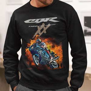 Op de afbeelding: Zwart sweatshirt met een motorfietsafbeelding en de tekst "CBR XX 1100 Super Blackbird" in zilver. De afbeelding toont een blauwe motorfiets en een rijder tegen een vurige achtergrond. Het sweatshirt heeft een ronde hals.