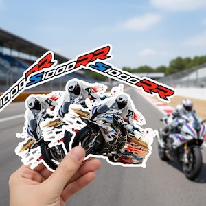 Könnte beinhalten: Eine Hand hält einen Aufkleberbogen mit Bildern eines BMW S1000RR Motorrads, das auf einer Rennstrecke fährt. Die Aufkleber haben einen weißen Hintergrund und den Text "S1000RR" in Rot, Weiß und Blau.