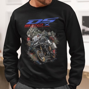 Pode incluir: Camisola preta com um gráfico de motocicleta e o texto "DS 900X" em azul e vermelho. O gráfico mostra uma motocicleta em uma paisagem montanhosa com efeitos de respingos de tinta coloridos.
