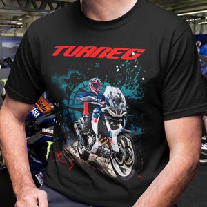 Pode incluir: Uma t-shirt preta de manga curta com uma estampa gráfica de um motociclista numa moto de aventura branca e azul. A palavra TUAREG está impressa em letras vermelhas grandes acima do condutor, sobre um design de bússola azul.