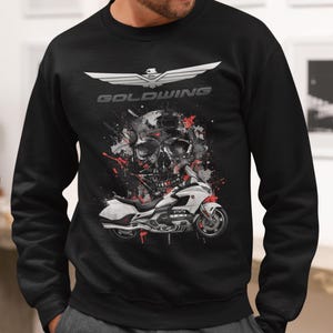 Pode incluir: Camisola preta com um gráfico de motocicleta e caveira. A palavra "GOLDWING" é impressa em branco acima da caveira. O design inclui respingos de tinta vermelha. Estilo casual.