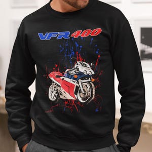 May include: Black sweatshirt featuring a motorcycle graphic with red, white, and blue accents. The text "VFR 400" is displayed above the motorcycle. The design includes paint splatter effects.