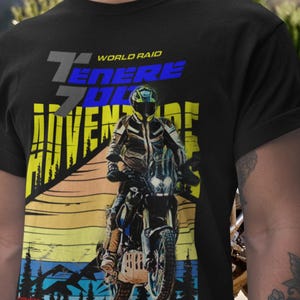 Pode incluir: Camiseta preta com um gráfico de um motociclista e o texto "World Raid Tenere 700 Adventure". O design apresenta um pôr do sol e uma paisagem montanhosa, com detalhes em azul, amarelo e cinza.