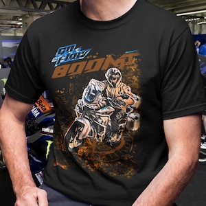 May include: Black t-shirt featuring a motorcycle graphic with a rider. The design includes the text "Go the Flow Boom!" in blue and orange. The image has a distressed, textured effect, suggesting speed and action.