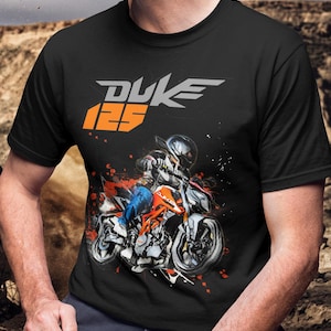 May include: Black t-shirt featuring a motorcycle graphic with the text "DUKE 125" in gray and orange. The graphic depicts a rider on an orange and white motorcycle with red paint splatter accents.