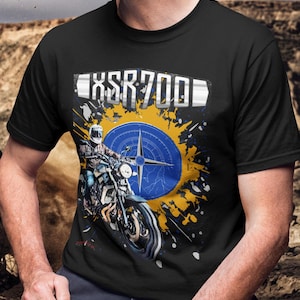 Pode incluir: Camiseta preta com um gráfico de motocicleta e o texto "XSR700". O design inclui uma bússola e um motociclista na moto. O fundo tem respingos de tinta amarelos e azuis.
