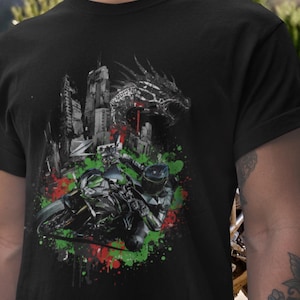 Op de afbeelding: Zwart t-shirt met een motorrijder ontwerp, tegen een stadsgezicht en een draak grafisch. De afbeelding bevat groene en rode verfspatten. De rijder draagt een helm. Het ontwerp is gedetailleerd en dynamisch.