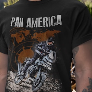 Könnte beinhalten: Schwarzes T-Shirt mit dem Text "PAN AMERICA" in Weiß. Das Shirt zeigt eine Grafik eines Motorradfahrers auf einer unbefestigten Straße, mit einem Kompass- und Kartendesign im Hintergrund. Das Design hat ein Vintage- und Abenteuer-Thema.
