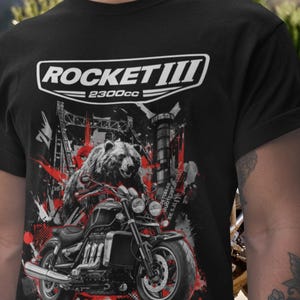 Może przedstawiać: Czarny t-shirt z grafiką przedstawiającą niedźwiedzia na motocyklu z białym napisem "ROCKET III 2300cc". Projekt zawiera czerwone akcenty i tło miejskie, tworząc odważny i dynamiczny obraz.