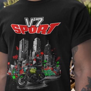 May include: Black t-shirt featuring a motorcycle graphic with the text "V7 SPORT" in red and white. The design includes a cityscape backdrop with graffiti art and a rider on a green motorcycle.