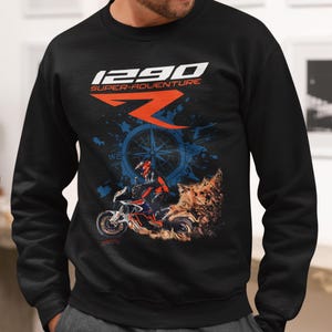 Pode incluir: Camisola preta com um gráfico de motocicleta e o texto "1290 SUPER-ADVENTURE". O design inclui uma bússola, um piloto e uma imagem estilizada de um animal. A camisola é de gola redonda.