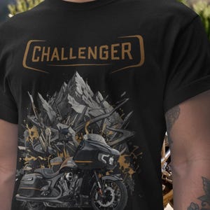 Könnte beinhalten: Schwarzes T-Shirt mit einem Motorrad-Motiv und dem Wort "CHALLENGER" in einem goldenen Rechteckrahmen. Das Motorraddesign enthält Berg- und abstrakte Elemente in Grau- und Goldtönen.