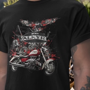 Peut inclure: T-shirt noir avec un motif de moto rouge et blanc. Le design comprend le mot "VALKYRIE" dans une bannière, des pièces de moteur et un aigle. Le t-shirt a un col rond.