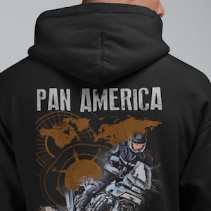 Pode incluir: Moletom preto com a inscrição "PAN AMERICA" em branco. O design apresenta um motociclista, uma bússola e um mapa-múndi em tons de marrom e laranja.