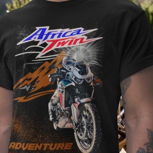 Może przedstawiać: Czarna koszulka z grafiką motocykla. Projekt zawiera napis "Africa Twin" w kolorze niebieskim i czerwonym, motocykl, góry i słowo "Adventure".