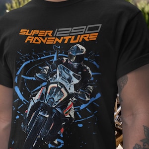 May include: Black t-shirt featuring a motorcycle graphic with the text "SUPER 1290 ADVENTURE" in orange and gray. The design includes a rider on a motorcycle with blue accents, perfect for motorcycle enthusiasts.