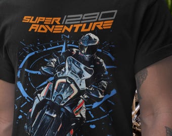 May include: A black t-shirt featuring a graphic of a motorcyclist riding a motorcycle. The text "SUPER 1290 ADVENTURE" is printed in orange and gray above the rider. A blue compass design is in the background behind the motorcycle.