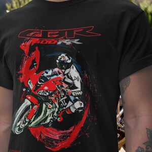 May include: Black t-shirt featuring a graphic of a red and white motorcycle with a rider. The text "CBR 600 RR" is displayed in red above the motorcycle. The shirt has a crew neck and short sleeves.