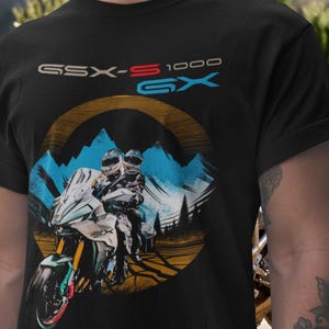 May include: Black t-shirt with a motorcycle graphic, including the text "GSX-S 1000 GX". The design features a motorcycle, riders, mountains, and a circular sun-like element. The shirt has a crew neck.