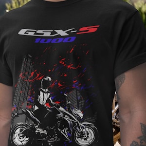 May include: Black t-shirt featuring a motorcycle graphic with the text "GSX-S 1000" in white, red, and blue. The design includes a cityscape backdrop and a rider on the motorcycle. The shirt is made of a dark fabric.