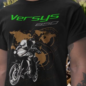 Pode incluir: T-shirt preto com um gráfico de motocicleta e o texto "Versys 650" em verde e cinza. O design inclui um mapa-múndi e uma bússola, sugerindo um tema de viagem ou aventura.