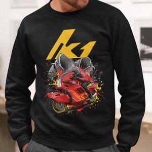 May include: Black sweatshirt featuring a graphic of a red motorcycle with a rider, set against a backdrop of yellow and red paint splatters. The sweatshirt also displays a large yellow "K1" logo.