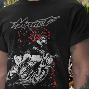Può includere: T-shirt nero con grafica di moto e la scritta "Hornet". Il design mostra un motociclista su una moto con schizzi rossi, creando un'estetica dinamica. Un ottimo regalo per gli appassionati di moto. Il design è stampato su una t-shirt nera.