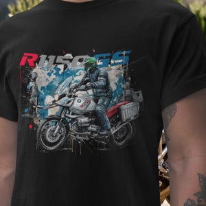 Pode incluir: Uma t-shirt preta com uma ilustração gráfica de uma pessoa a conduzir uma mota BMW GS prateada. O design inclui um fundo de mapa-mundo azul, uma rosa dos ventos e o texto R1150GS em vermelho e azul na parte superior.