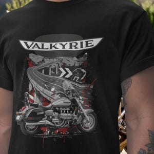 Peut inclure: T-shirt noir avec un motif de moto et le mot "VALKYRIE" au-dessus. Le design comprend une route sinueuse, un pont et des éclaboussures de peinture rouge, créant une esthétique dynamique et audacieuse.