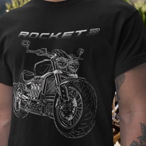 Puede incluir: Camiseta negra con un dibujo lineal blanco de una motocicleta y el texto plateado "ROCKET 3". El diseño de la motocicleta incluye los faros, las ruedas y el motor. La camiseta es de cuello redondo.
