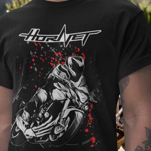 May include: Black t-shirt featuring a motorcycle graphic with a rider in grayscale. The word "Hornet" is displayed in white, with a stylized lightning bolt design. Red splatter accents the design.