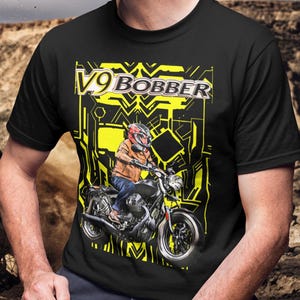 Puede incluir: Camiseta negra con un gráfico amarillo que presenta una motocicleta y el texto "V9 BOBBER". El diseño incluye un piloto con casco y chaqueta en la motocicleta. El fondo tiene un patrón geométrico.