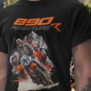 Könnte beinhalten: Schwarzes T-Shirt mit einer orangefarbenen und grauen Grafik. Die Grafik zeigt den Text "890 Adventure" und eine Illustration eines Motorradfahrers auf einem Dirtbike. Das Design beinhaltet einen Kompass und eine Berglandschaft.