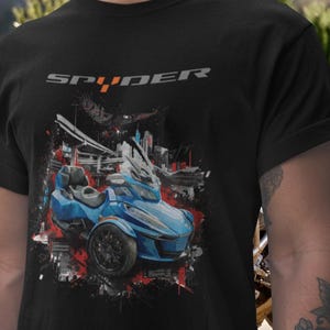 Puede incluir: Camiseta negra con un gráfico de una motocicleta Spyder azul de tres ruedas. El diseño incluye la palabra "SPYDER" en gris, con un paisaje urbano y un efecto de salpicaduras de pintura roja y blanca. La motocicleta es el punto focal.