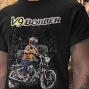 Puede incluir: Camiseta negra con un gráfico de motocicleta y el texto "V9 BOBBER". El gráfico muestra a una persona en una moto, con casco y chaqueta. El texto es amarillo y blanco.