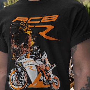 Puede incluir: Camiseta negra con un gráfico de motocicleta naranja y blanco y el texto "RC8 R". El diseño incluye una calavera y llamas. La camiseta está hecha de un material suave.