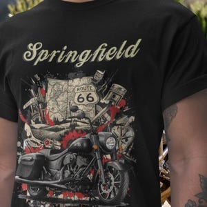 May include: Black t-shirt featuring a motorcycle graphic with the text "Springfield" above and "Route 66" on a map. The design includes engine parts and red accents, creating a vintage, biker theme.