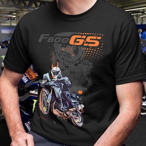 以下が含まれることがあります： オレンジ色の「F800 GS」の文字とバイクのグラフィックが特徴の黒いTシャツ。デザインは、グレーとオレンジの配色で、バイクに乗ったライダーを描いています。このTシャツはおそらく綿または綿混紡で作られています。
