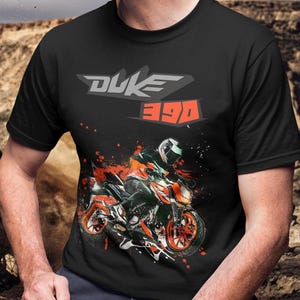 May include: A black short-sleeve t-shirt featuring a graphic print of a motorcycle rider on an orange and black motorcycle. The text DUKE 390 is printed in grey and orange above the motorcycle graphic, which includes orange paint splatter effects.