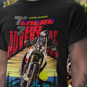 Pode incluir: Camiseta preta com um gráfico de motocicleta e o texto "Ténéré 700 Adventure" em vermelho e branco. O design apresenta um motociclista em uma moto contra um pôr do sol, com as palavras "World Raid" acima.