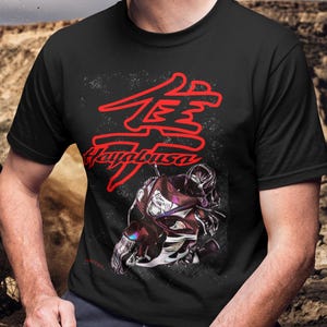 Op de afbeelding: Zwart T-shirt met een rode afbeelding van het woord "Hayabusa" in Japanse karakters en een motorfiets. Het ontwerp staat tegen een gespikkelde achtergrond. Het shirt heeft korte mouwen.