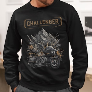 Könnte beinhalten: Schwarzes Sweatshirt mit einem Motorrad-Motiv und dem Wort "CHALLENGER" in einem goldenen Rechteck. Das Motorrad ist schwarz und grau mit detailliertem Design. Der Hintergrund zeigt eine bergähnliche Struktur.