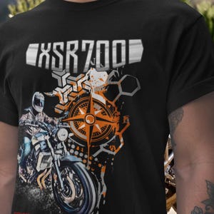 Pode incluir: Camiseta preta com um gráfico de motocicleta e o texto "XSR700". O design inclui uma bússola, formas geométricas e um motociclista. A camiseta tem gola redonda.