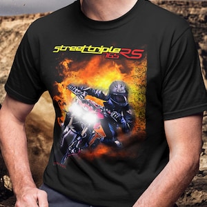 May include: Black t-shirt featuring a motorcycle graphic with a rider in a helmet, set against a fiery background. The text "street triple RS 165" is displayed in yellow and red. The shirt is a classic crew neck.