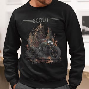 Pode incluir: Camisola preta com um gráfico de motocicleta e a palavra "SCOUT". O design inclui um lobo e uma motocicleta em tons de verde, castanho e preto. A camisola é de gola redonda.
