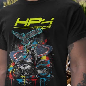 Op de afbeelding: Zwart T-shirt met een motorrace-ontwerp. De afbeelding toont een motorfiets, een racebaan en een adelaar, met de tekst "HP4 RACE" in geel. Kleurrijke verfspatten dragen bij aan het dynamische gevoel.