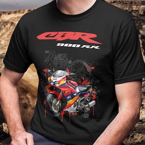 Pode incluir: T-shirt preto com um gráfico de uma moto vermelha, azul e amarela e o texto "CBR 900RR" em vermelho. O design inclui respingos de tinta vermelha. A camiseta tem um decote redondo clássico. O gráfico da moto é detalhado.