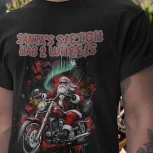 Puede incluir: Camiseta negra con un diseño de Papá Noel montado en una moto roja. El texto "SANTA'S SLEIGH HAS 2 WHEELS" está en rojo y blanco. Papá Noel lleva un traje rojo y gafas de sol, con un saco de regalos.